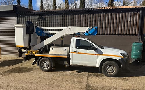 White pickup with a CPL platform on the rear, boom folded back, parked outside a shed