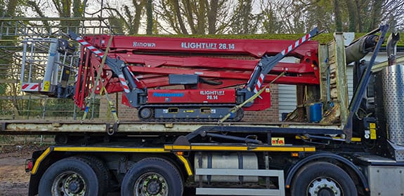 Red tracked spider platform with booms folded, loaded on the back of a flatbed lorry
